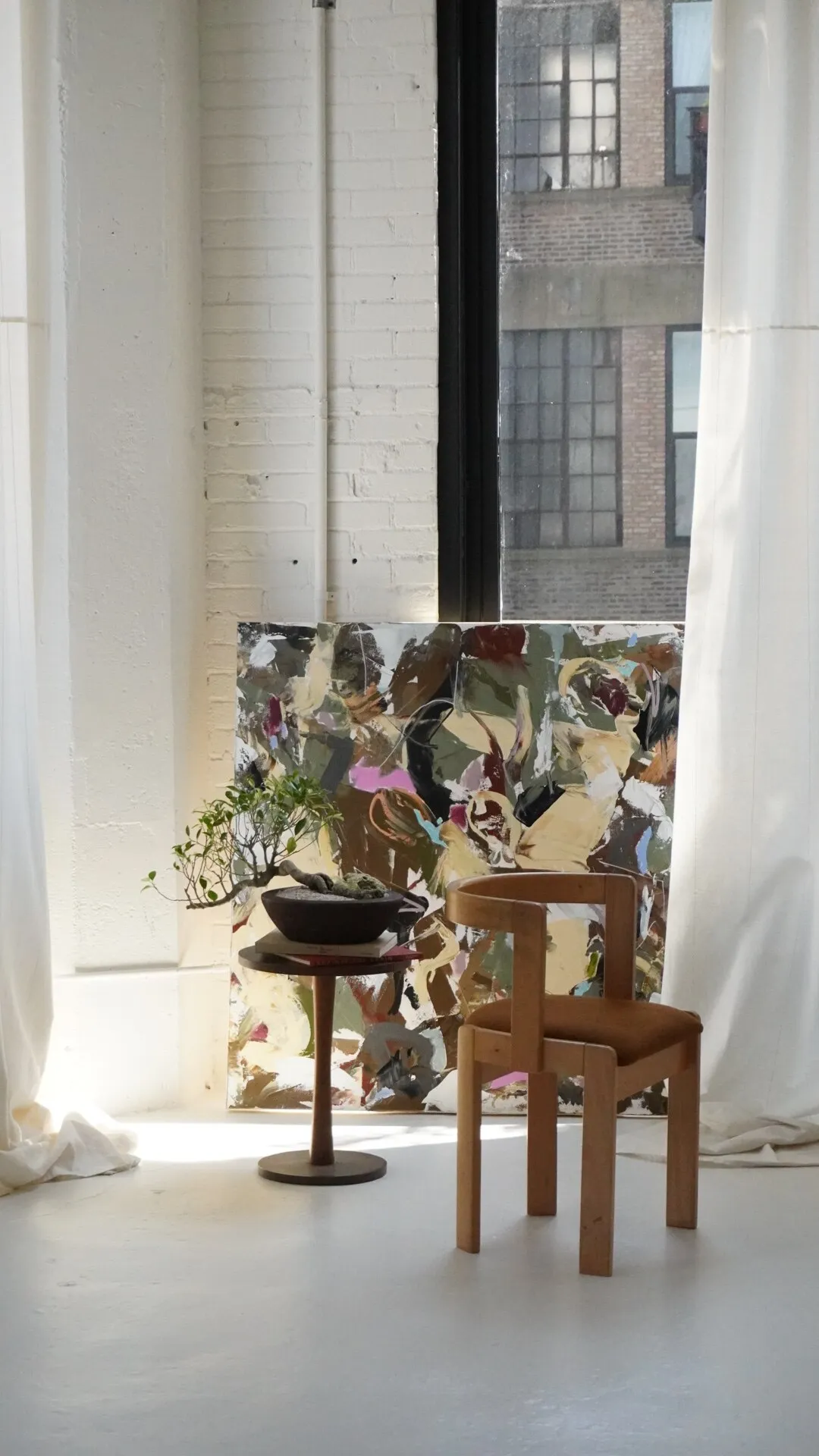 A cascading ficus bonsai in a dark bowl on a pedestal table, staged against a large abstract painting in a loft with factory windows.
